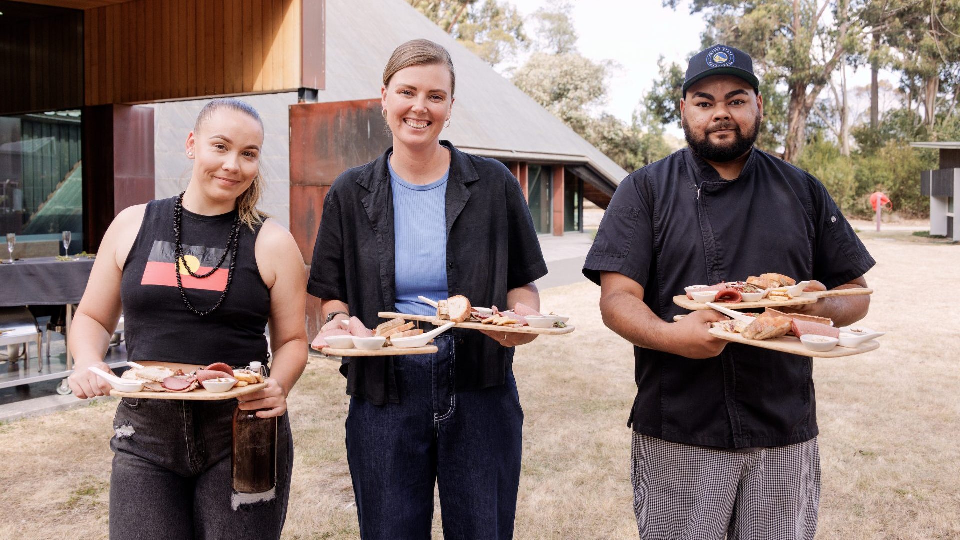 Two women and a man hold plates of food and smile to camera