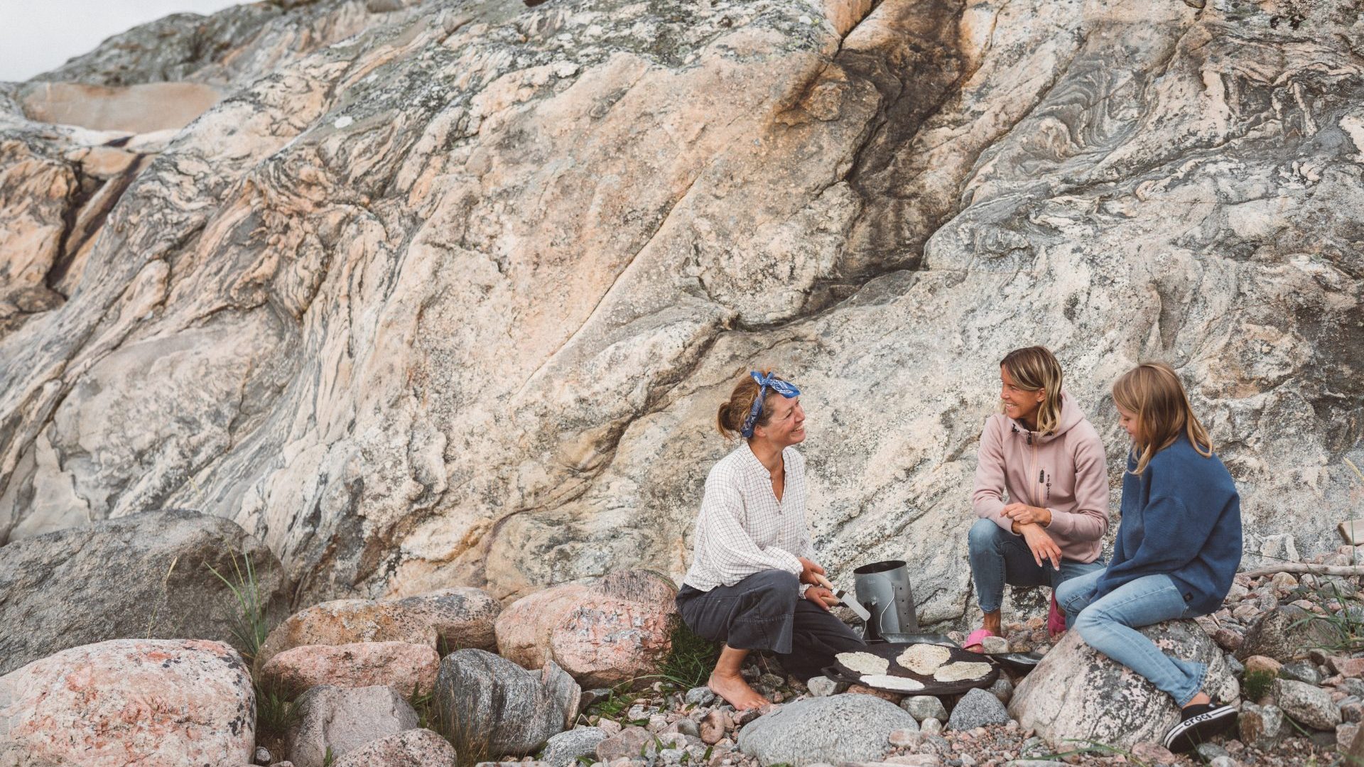 Three people gather beside sandy coastal rocks cooking over a camp stovetop.