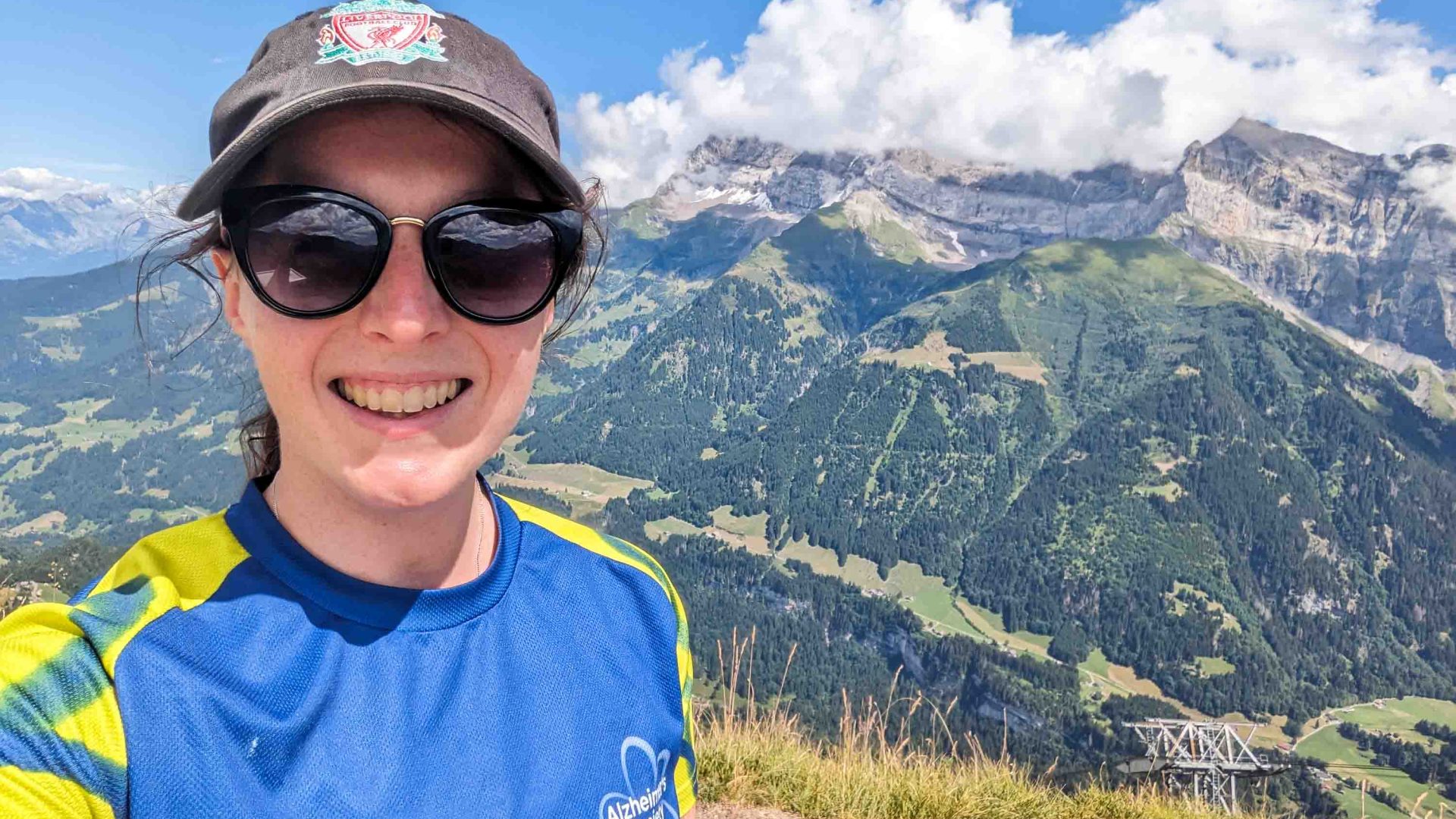 A woman in a hat and sunglasses smiles to camera. She stands in front of mountains.