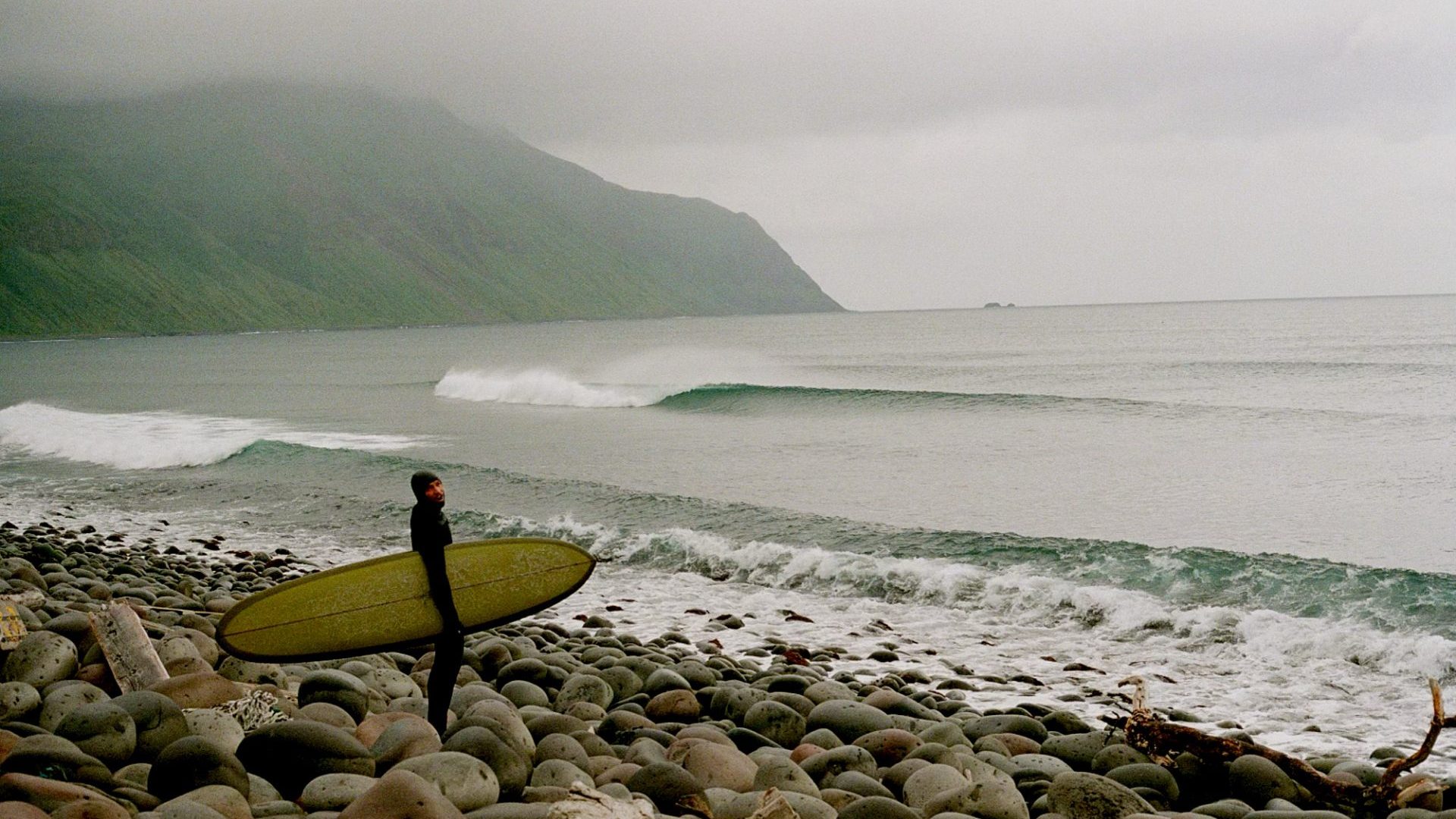 Surfer wearing full body wet suit and hood smiles at camera from rocky beach