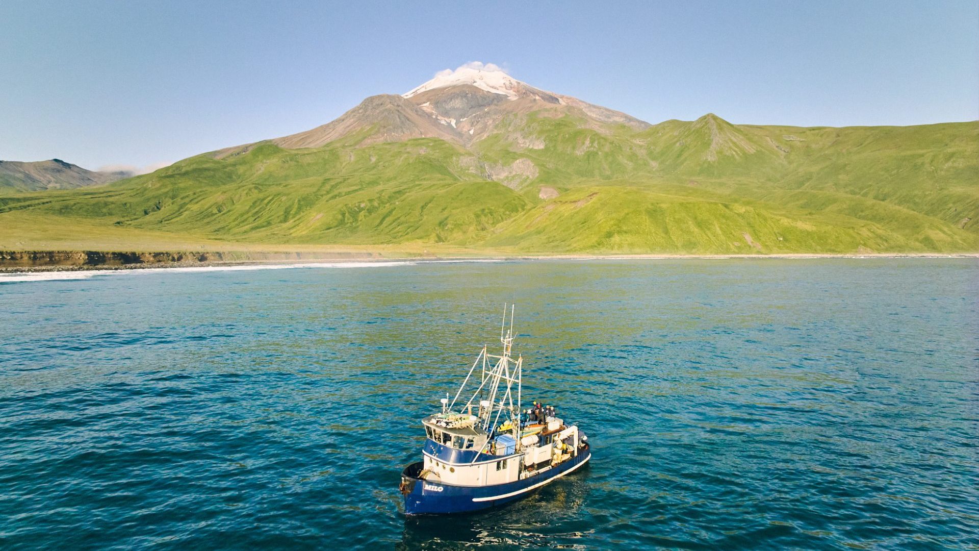 Boat in blue water against a green mountain with snow capped volcano