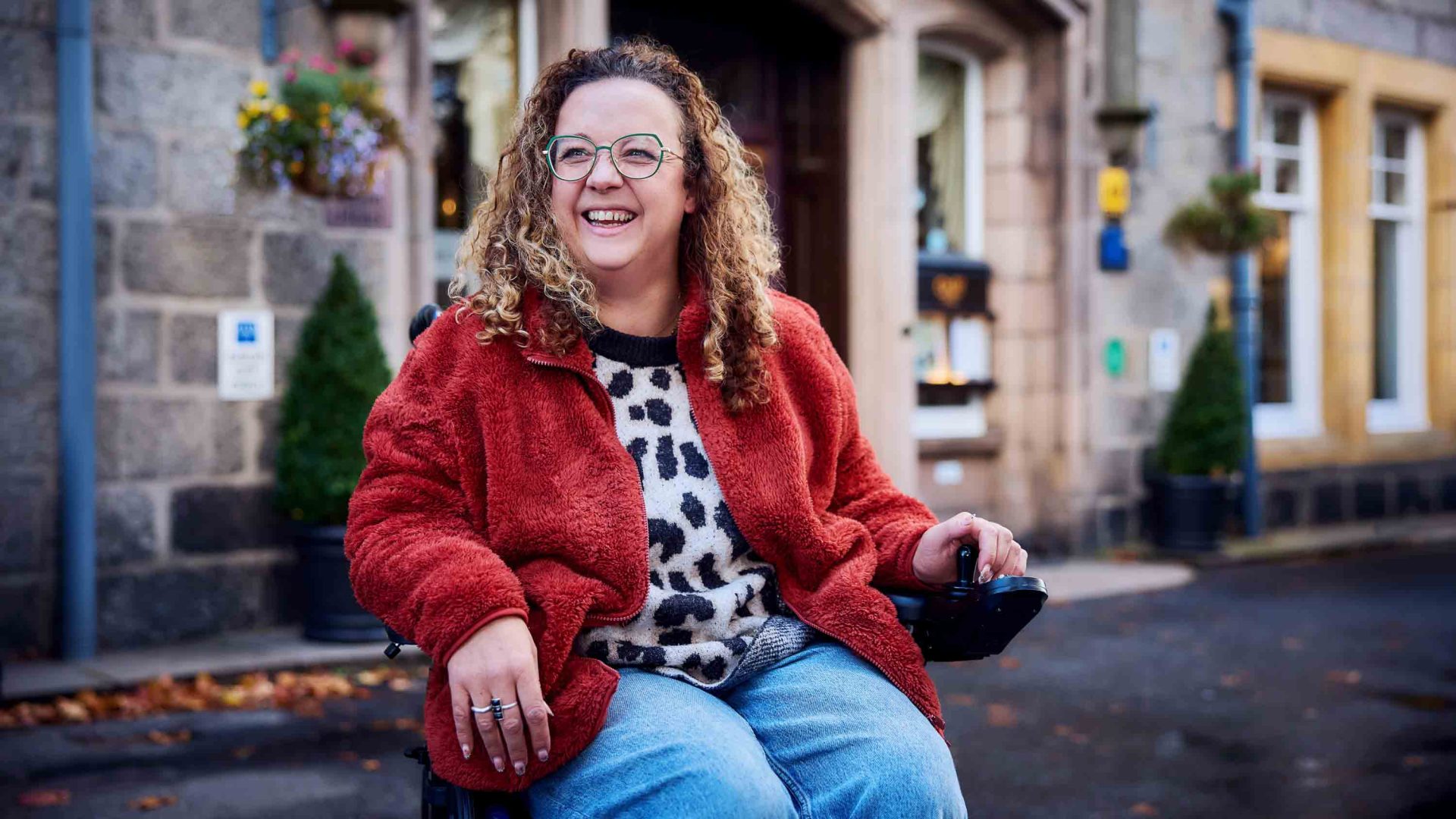 A woman in a wheelchair with long curly blonde hair smiles as she sits outside a stone hotel.