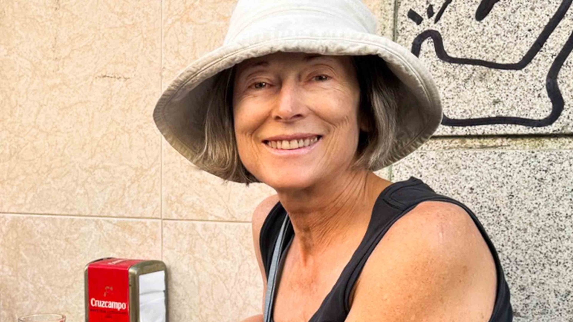 A woman in a white hat who is seated at a table smiles to camera.