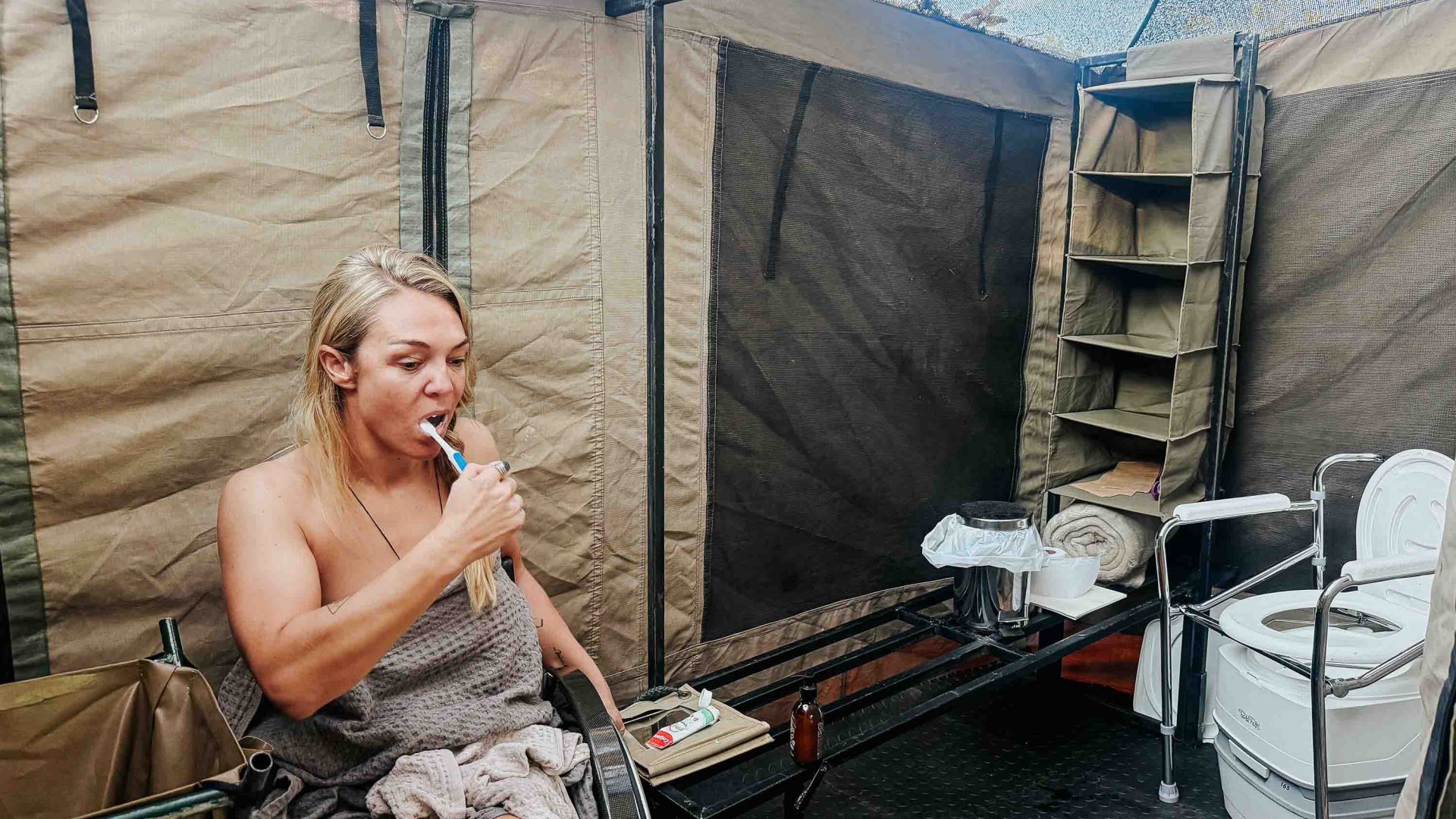 A woman brushes her teeth with a camp toilet and shower behind her. She is in a heelchair.