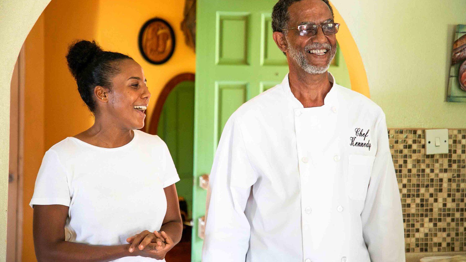 A woman and a man smile. They are ina room with a green door and look away from camera.