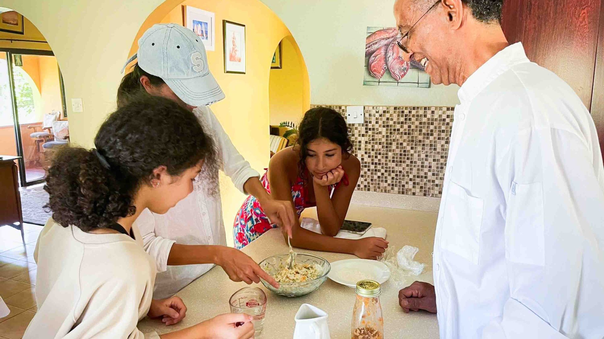 A group of three people round a mixing bowl in a kitchen while a chef looks on smiling.