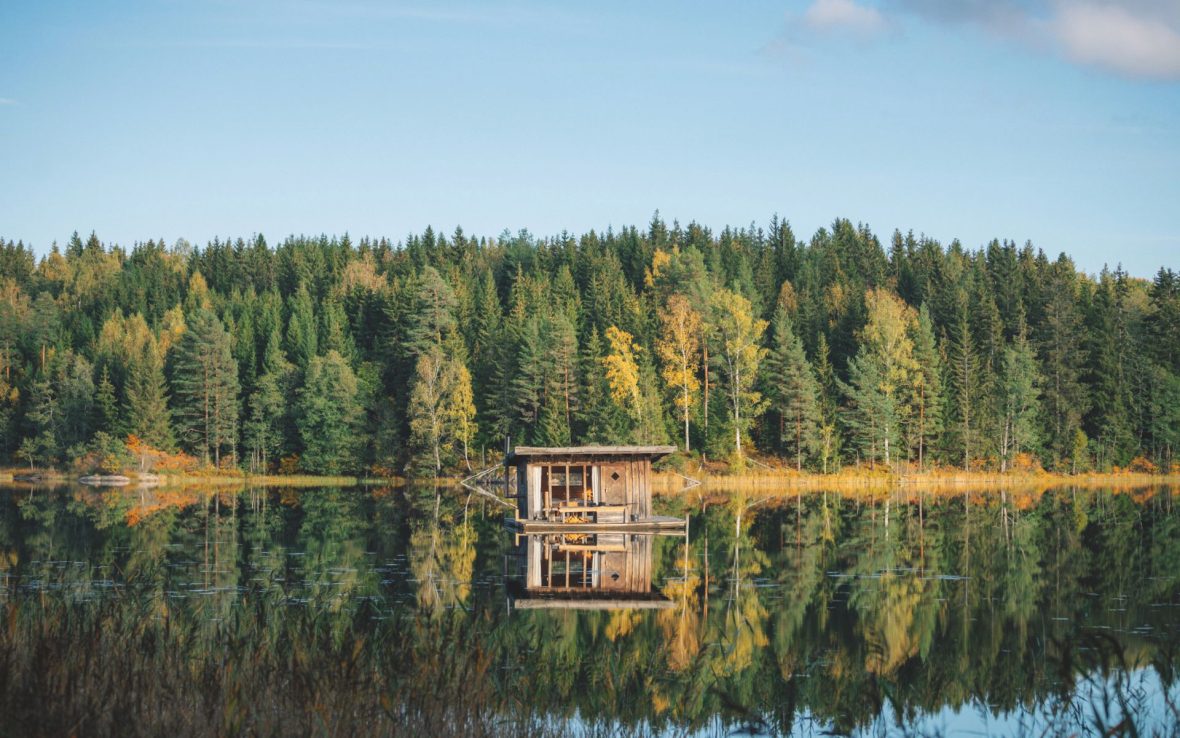 View of a small wooden cabin from across a lake in Sweden, with an evergreen forest in the background.