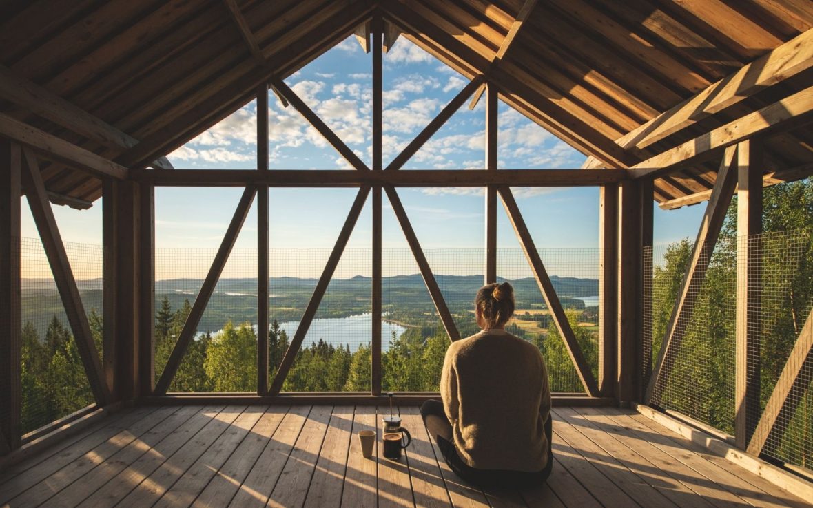A traveler sits facing away from the camera, looking out over a forest view from a wooden cabin in Sweden with large windows. It's very peaceful.