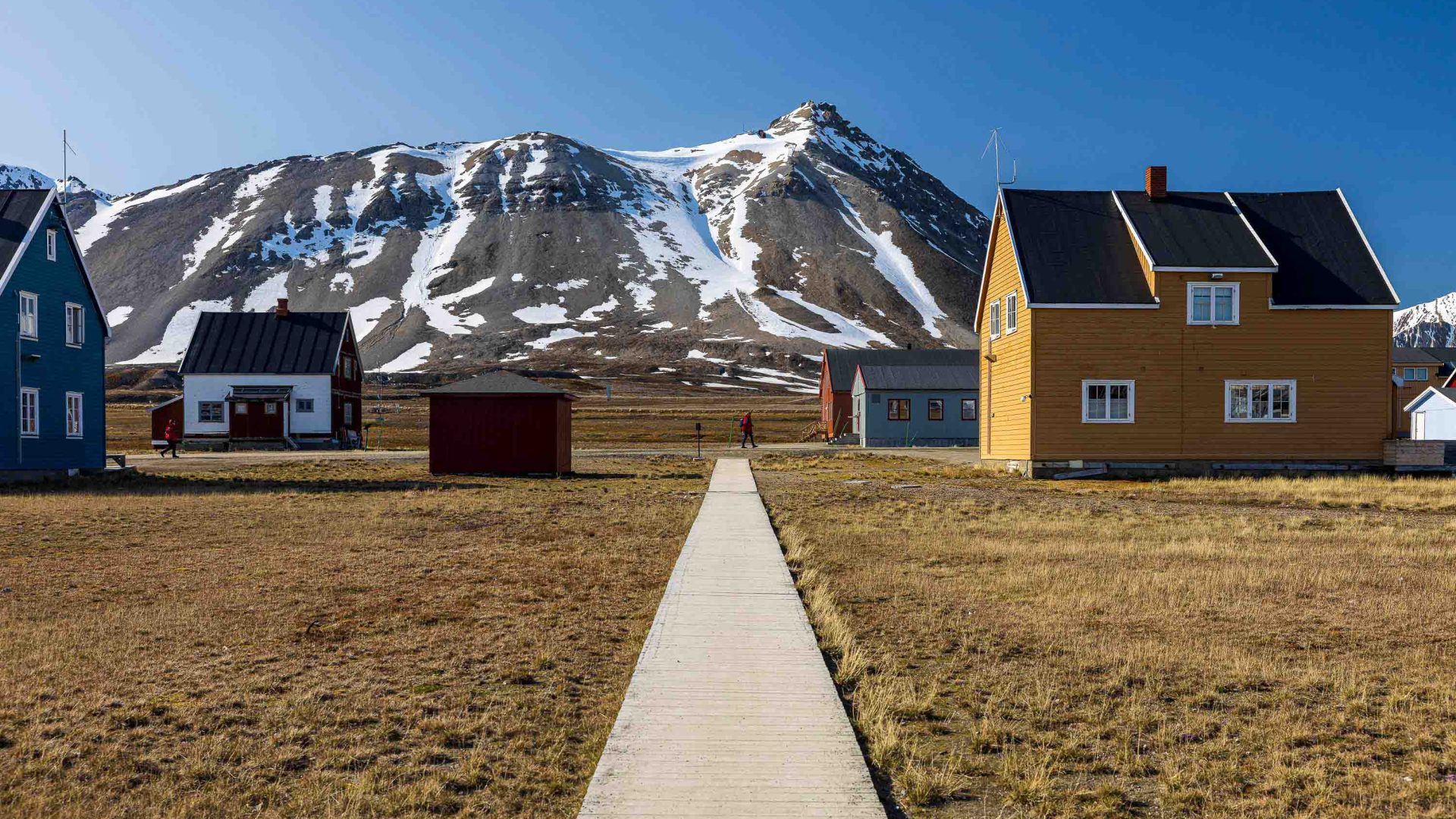 A path leads up to a few colourful buildings against a backdrop of mountains.