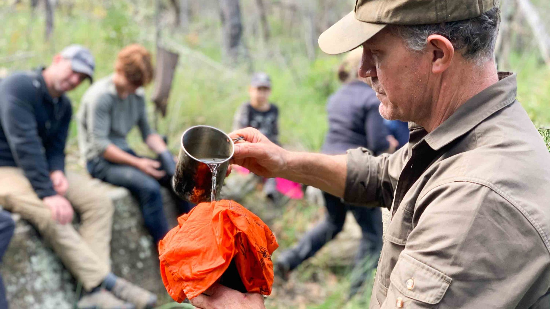 A man pours water over an orange bandana while a group looks on.