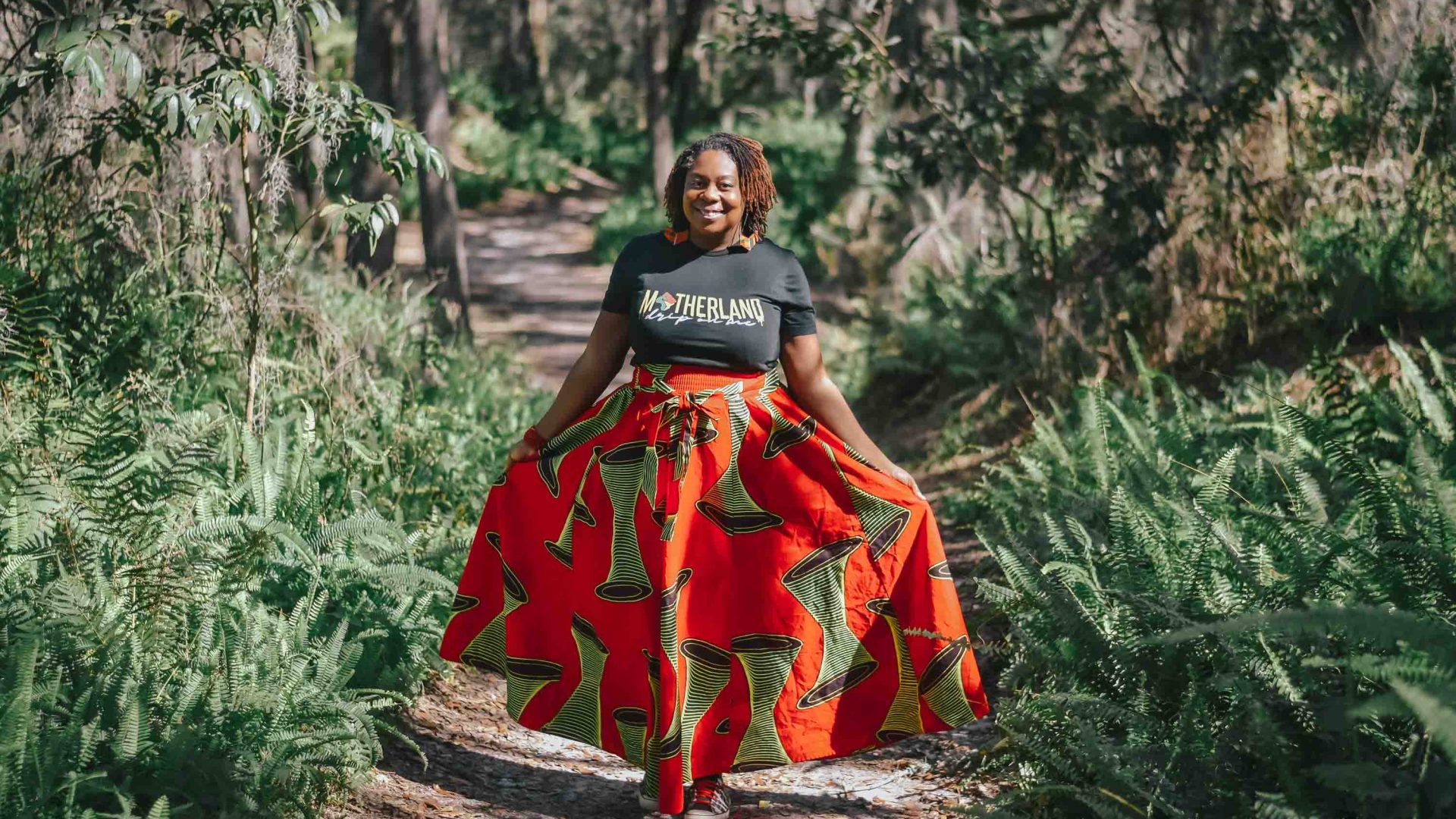 A woman smiles to camera wearing a black 'Motherland' top and a bright red skirt