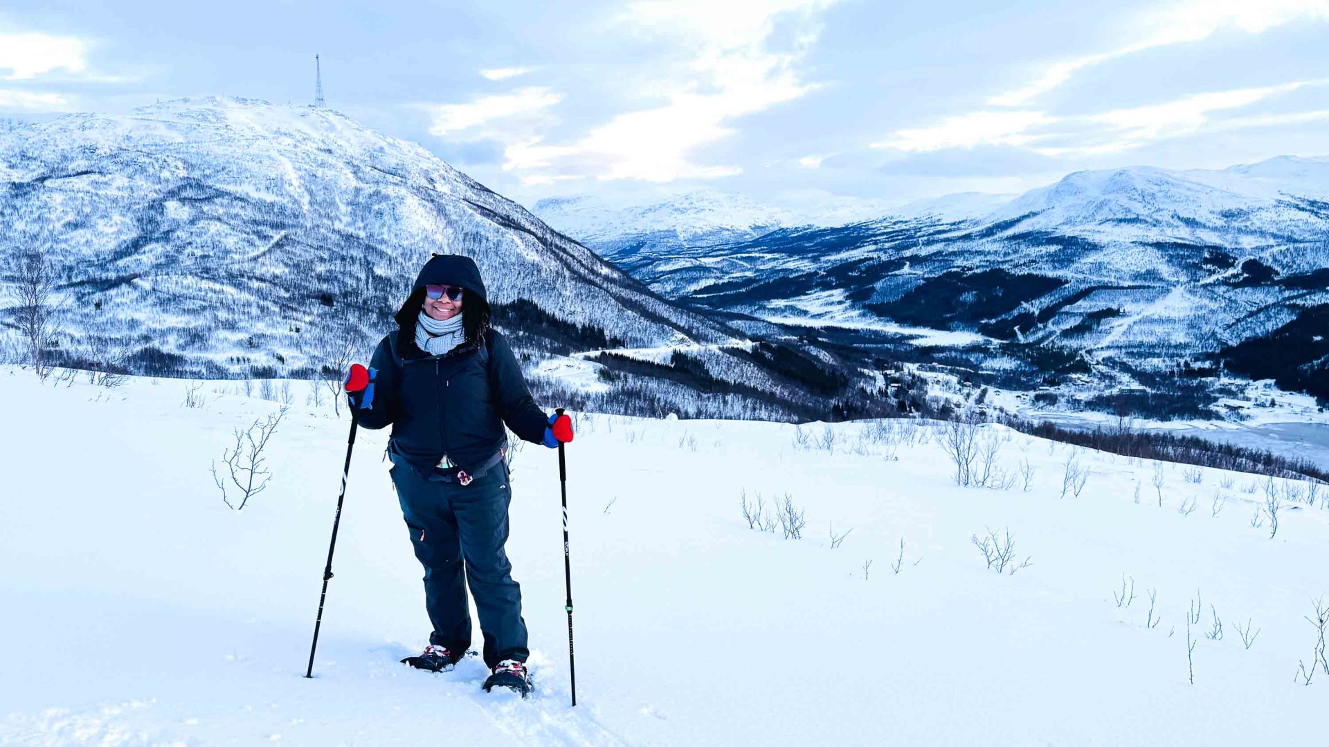A woman in snow shoes stands and smiles against a snowy mountainous backdrop.