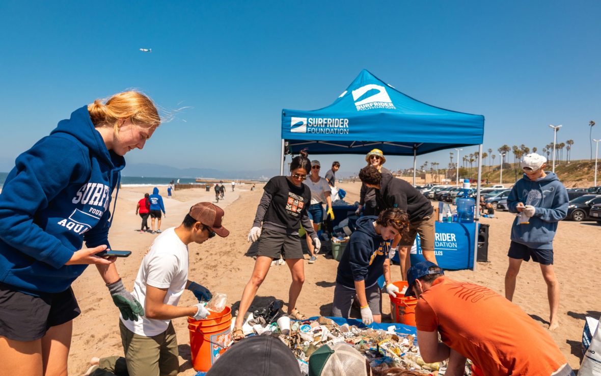Team on beach stand below tent with 'Surfrider Foundation' printed on it. They're picking up trash from the beach.