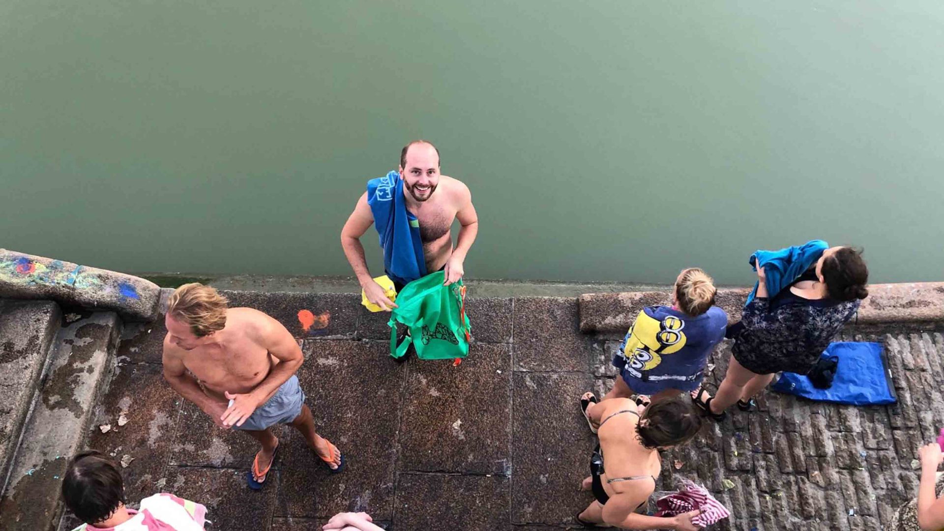 A male swimmer on a platform looks up and smiles, while others get ready for a dip