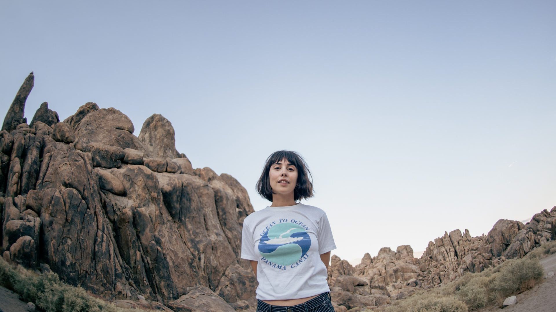 Woman wearing white t-shirt stands beneath tall rocks