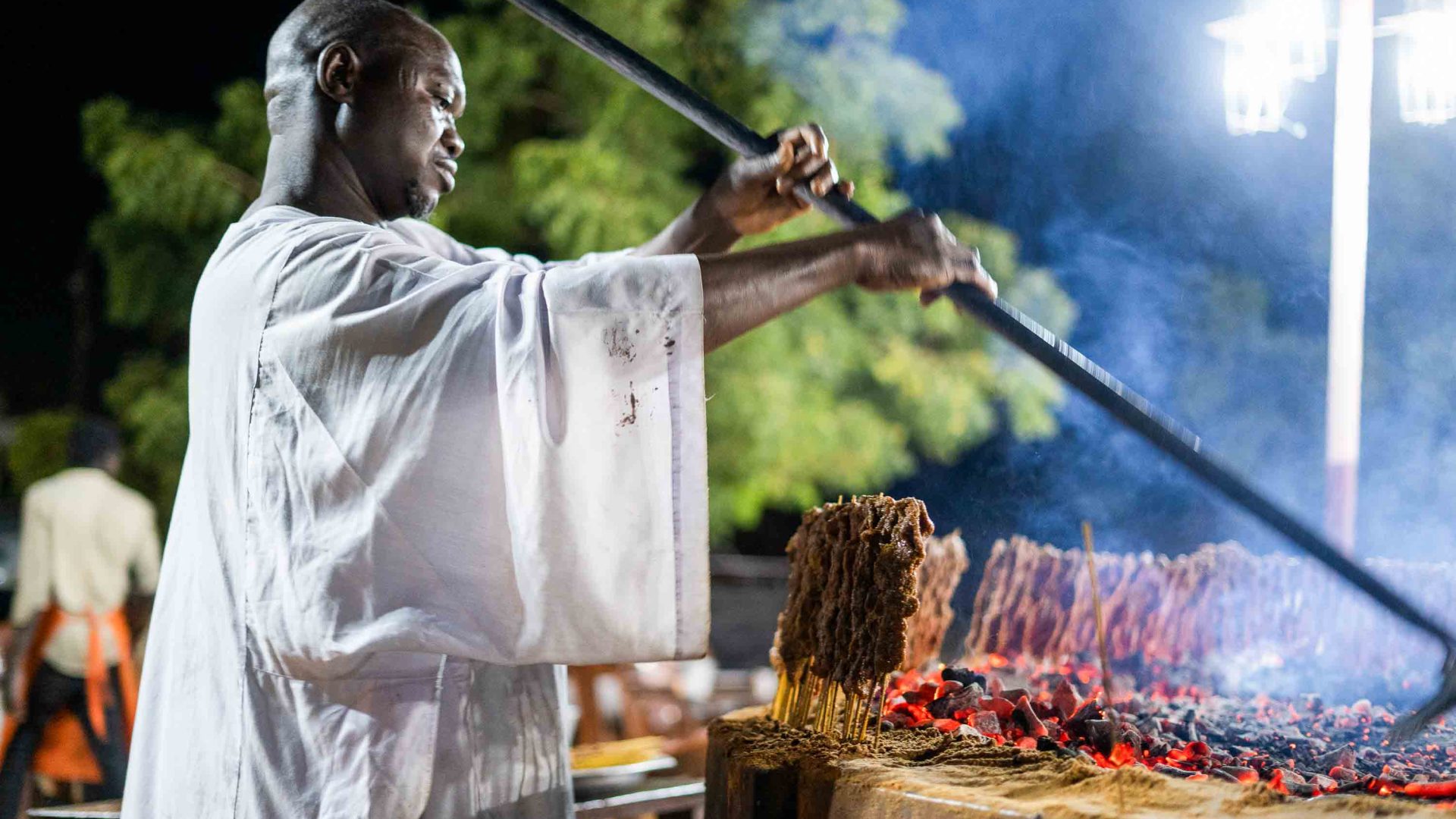 A man moves coals that are cooking meat on skewers.