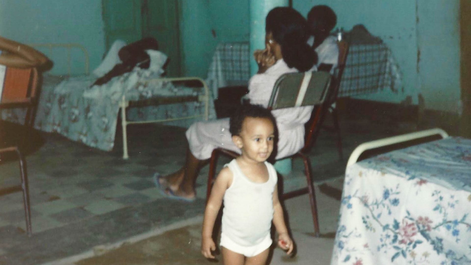 A toddler walks through a house where adults are seated, watching TV.