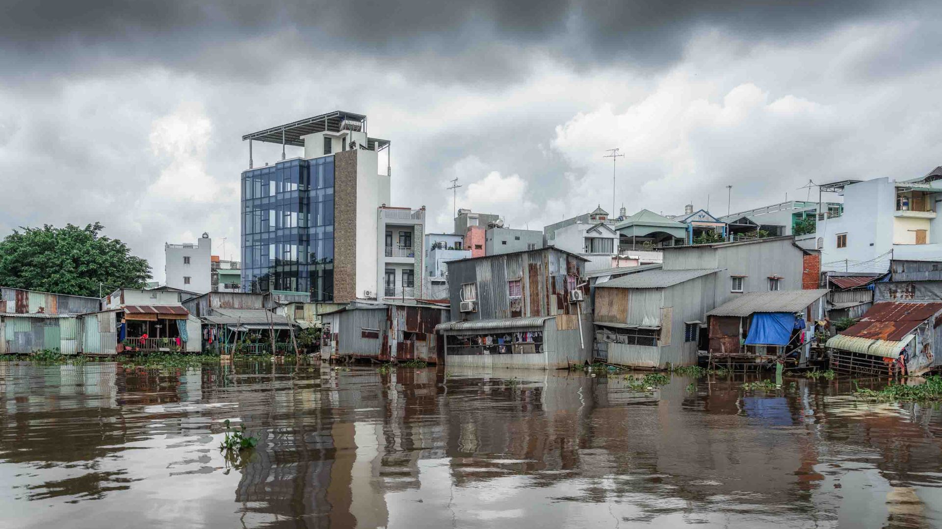 Houses sit in the brown water of the river. A grey cloudy sky is above them.