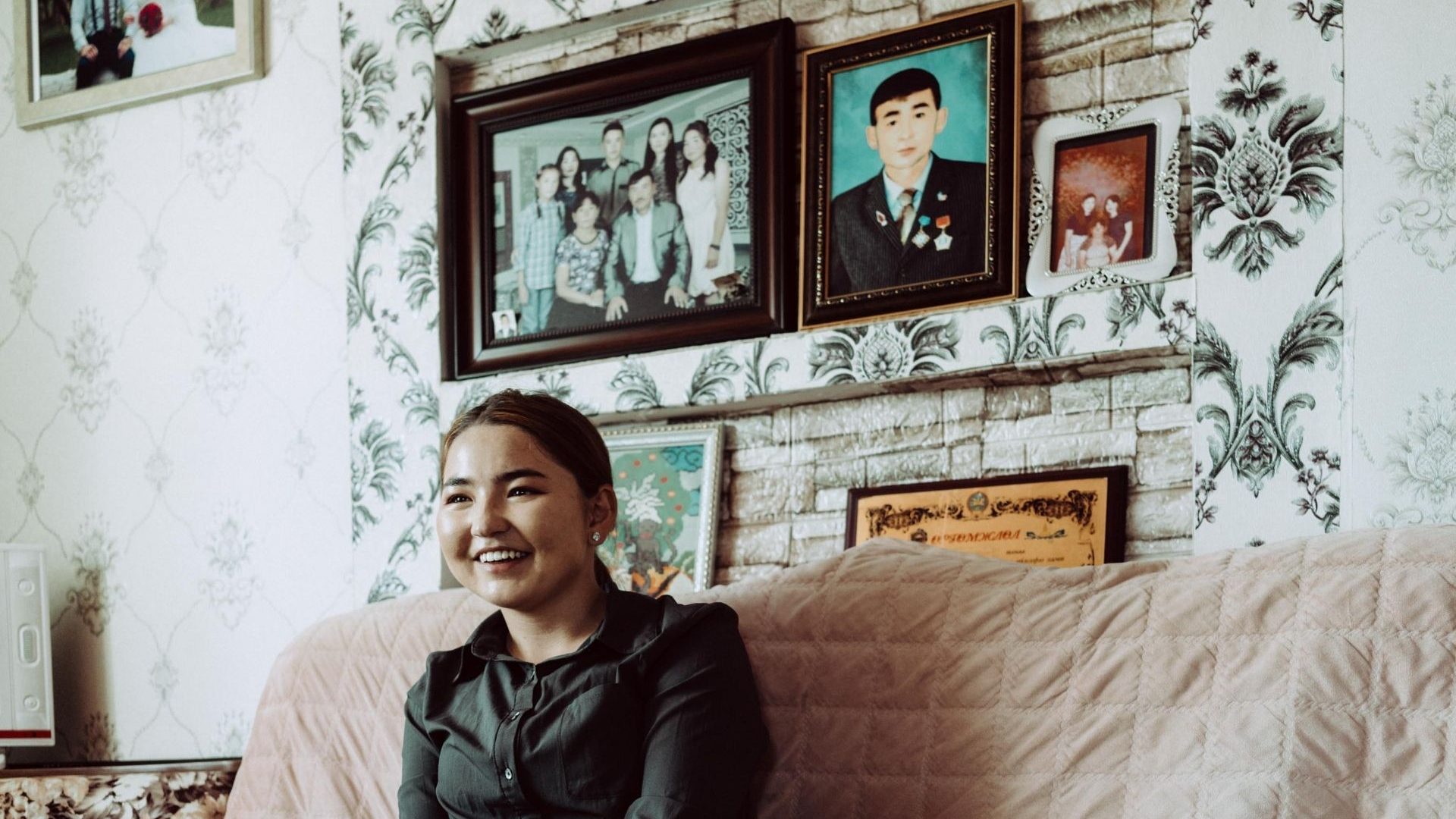 A woman in a black shirt with dark hair smiles to camera, sat on a sofa in her home with family photos on the wall.